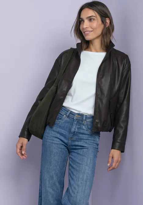 Frau in schwarzer Lederjacke, weißem Shirt und blauen Jeans vor lila Hintergrund.