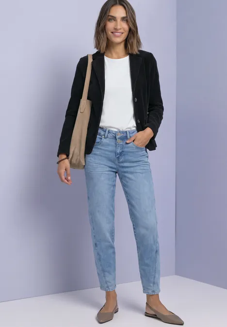 Frau vor lila-grauem Hintergrund in schwarzer Jacke, weißem Shirt, blauen Jeans und flachen Schuhen, mit Tasche.