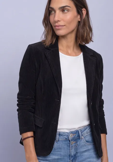 Frau mit schwarzem Blazer, weißem Top und Jeans steht vor neutralem Hintergrund.