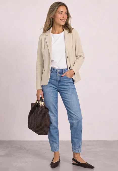 Frau in beigem Blazer, weißem T-Shirt, blauen Jeans und schwarzen Ballerinas mit dunkler Handtasche.