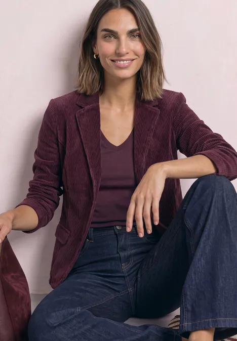 Frau sitzt lächelnd, trägt ein weinrotes Sakko, dunkles Top und weite Jeans vor hellem Hintergrund.