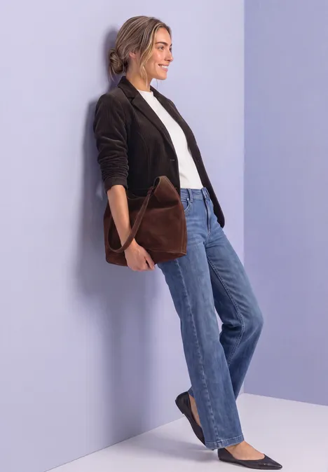 Une femme appuyée contre un mur pastel porte une veste noire, un top blanc, un jean bleu et un sac marron.