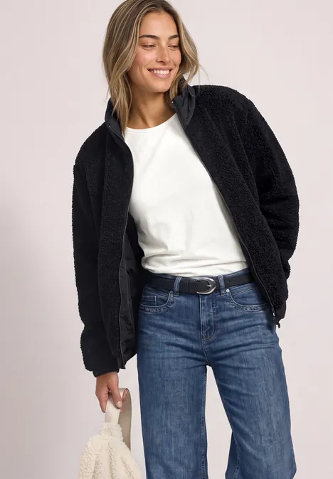 Frau in schwarzer Teddyjacke, weißem T-Shirt und Jeans, lächelt und hält ein Stoffstück.