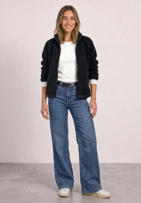Eine Frau trägt eine schwarze Teddyjacke, weißen Pullover, weite Jeans und weiße Sneakers vor hellgrauem Hintergrund.