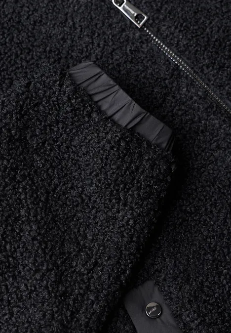 Nahaufnahme eines schwarzen Teddy-Fleece-Kleidungsstücks mit Reißverschluss, Raffung und Knopf.