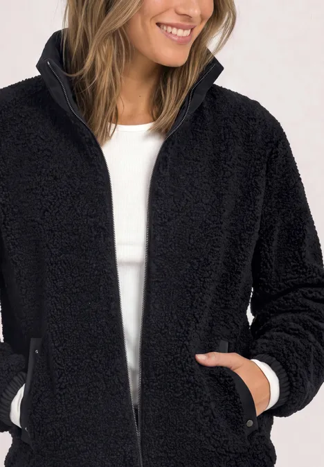 Eine Frau trägt eine flauschige schwarze Teddy-Fleecejacke mit Reißverschluss und lächelt.