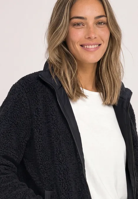 Eine lächelnde Frau in schwarzer Fleecejacke und weißem T-Shirt vor hellem Hintergrund.