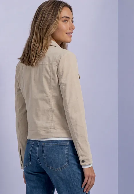 Eine Frau in beiger Jacke und blauen Jeans, seitlich stehend, lächelnd vor hellem Hintergrund.