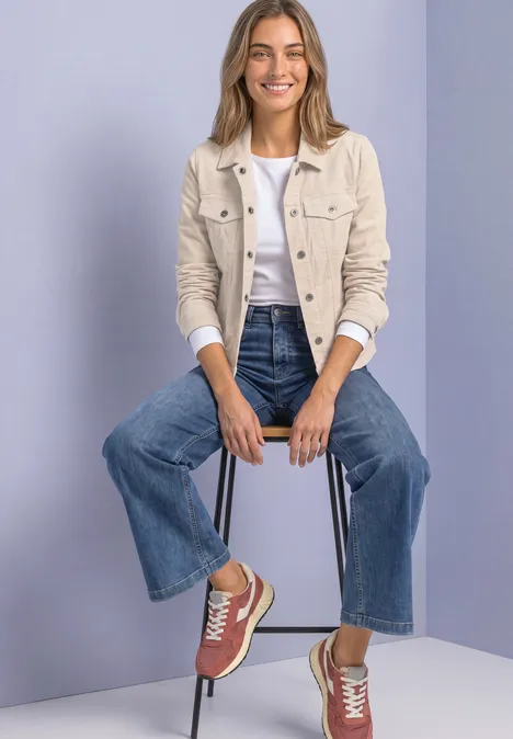 Eine Frau sitzt auf Hocker in Jeans, weißem Shirt, beiger Jacke und roten Sneakern vor violettem Hintergrund.