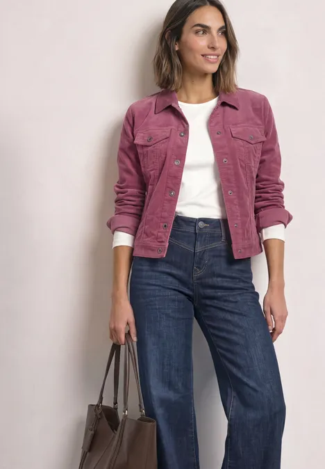 Frau trägt rosa Cordjacke, weißes Shirt, blaue Jeans und hält eine braune Tasche in der Hand.
