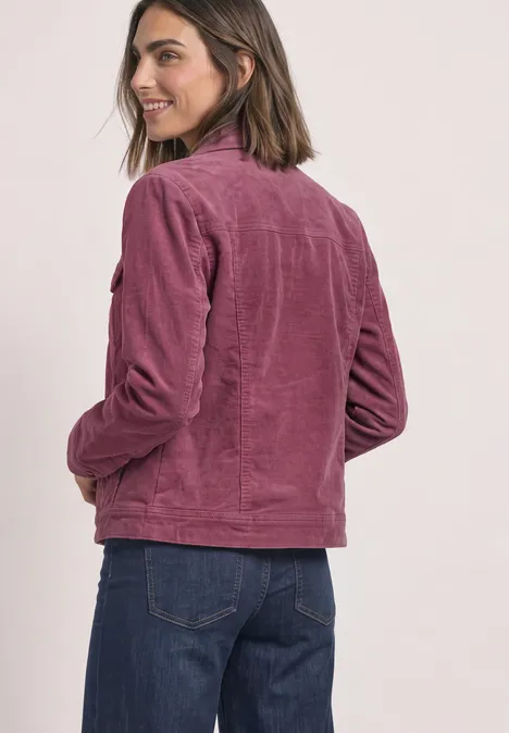 Junge Frau mit schulterlangen Haaren, trägt rosafarbene Cordjacke und blaue Jeans, lächelnd seitlich zur Kamera gewandt.