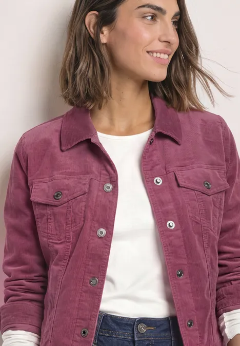 Frau in pinker Cordjacke mit weißem Shirt und Jeans lächelt seitlich.