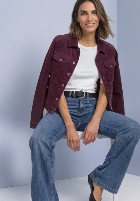 Frau sitzt lässig in weitem Jeans, weißem Shirt und Burgunderjacke vor hellgrauem Hintergrund.