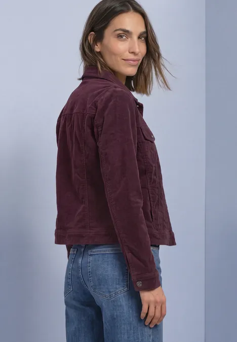 Frau in weinroter Cordjacke und blauen Jeans, steht seitlich und blickt zur Kamera vor schlichtem Hintergrund.