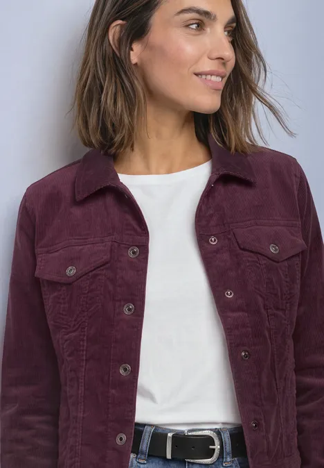 Frau trägt weiße Bluse unter lila Cordjacke mit Taschen und schwarzem Gürtel, lächelt seitlich.