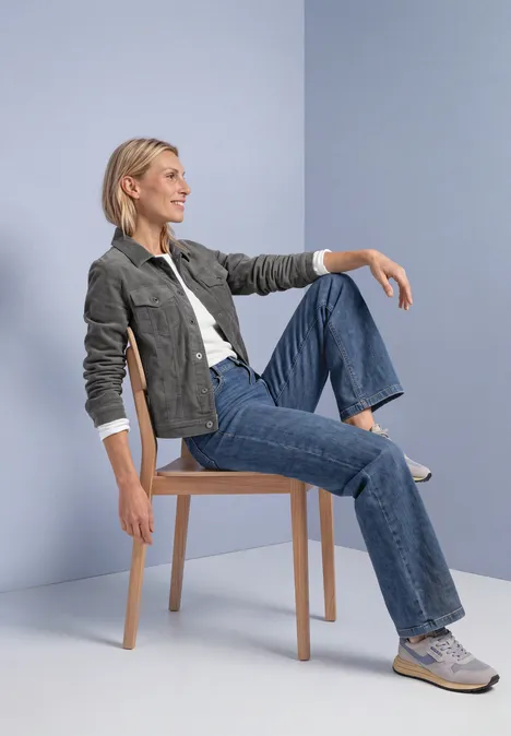 Une femme blonde est assise détendue sur un tabouret, porte un jean, baskets blanches et veste grise sur fond bleu.