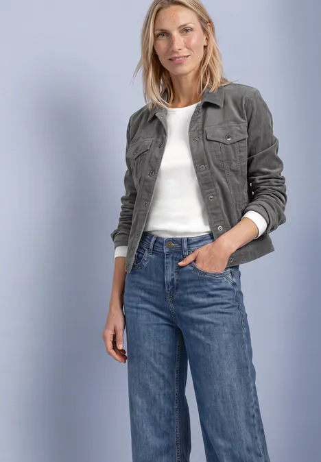 Femme en veste en velours côtelé vert foncé, haut blanc et jean bleu, souriante, main dans la poche.