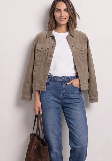 Frau trägt weißes T-Shirt, hellbraune Jacke, blaue Jeans und hält eine braune Tasche.