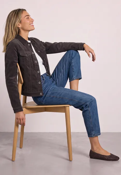 Femme assise décontractée sur une chaise, penchée en arrière, un genou replié, en jean, veste et chaussures plates.