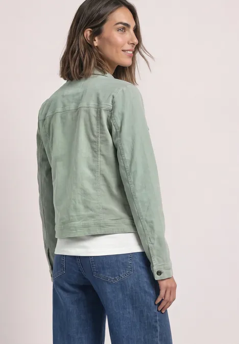 Femme de dos en veste verte et jean, regard par-dessus l'épaule sur fond épuré.