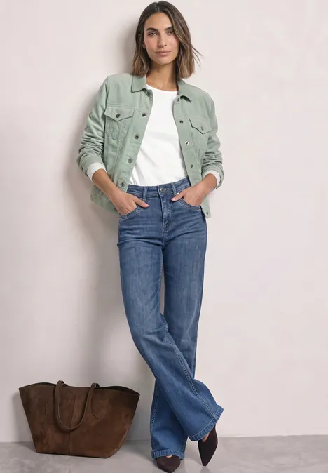 Femme en veste verte, t-shirt blanc et jean bleu, debout près d’un grand sac marron sur un fond clair.