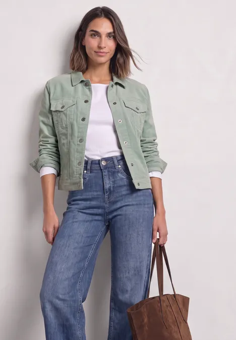 Femme en veste verte en velours, top blanc et jean, tenant un sac marron.
