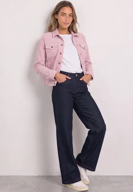 Femme en veste en jean rose, t-shirt blanc et jean large foncé, baskets blanches, fond clair.