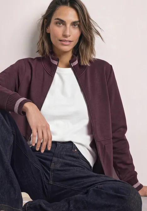 Veste de sport à col montant et détails en maille rustic rouge Frau mit kastanienfarbenem Blouson, weißem T-Shirt und Jeans sitzt lässig und blickt selbstbewusst.