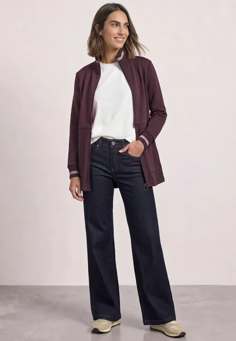 Veste de sport à col montant et détails en maille rustic rouge Frau in weißem Shirt, bordeauxfarbener Jacke und weiten, dunklen Jeans, sportliche Sneaker.