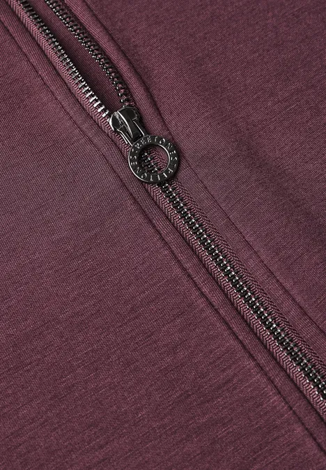Veste de sport à col montant et détails en maille rustic rouge Nahaufnahme eines schwarzen Reißverschlusses auf burgundrotem Stoff.