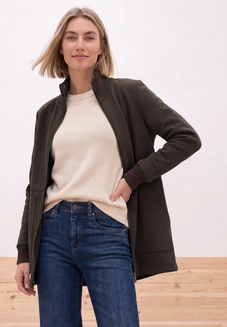 Vrouw in zwarte cardigan, beige shirt en jeans leunt nonchalant tegen lichte achtergrond.