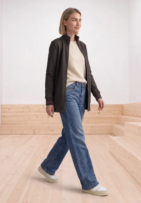 Jonge vrouw in zwarte jas, beige trui, blauwe jeans en witte sneakers loopt door een lichte ruimte.