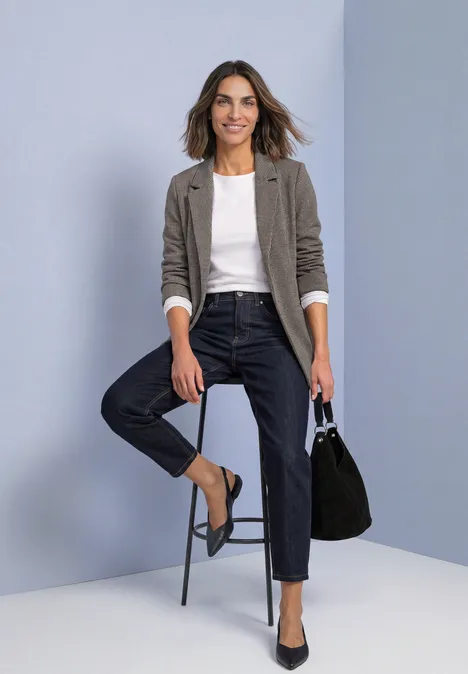 Frau im grauen Blazer, weißem T-Shirt und dunklen Jeans sitzt auf einem Hocker und hält eine schwarze Handtasche