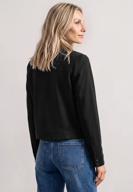 Een vrouw met de rug naar de camera kijkt glimlachend naar rechts, draagt een zwart shirt en blauwe jeans.