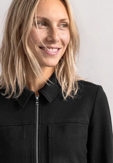 Close-up van een glimlachende blonde vrouw in een zwarte jas, die naar rechts kijkt.
