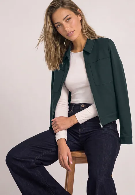 Vrouw zit ontspannen op krukje, draagt donkergroene jas, licht shirt en zwarte jeans.
