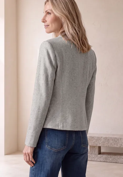 Frau von hinten in grauem Blazer und blauen Jeans vor hellem Hintergrund.