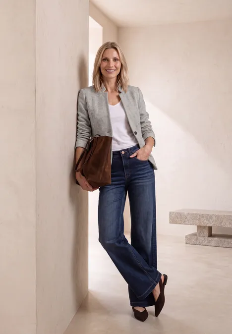Eine blonde Frau lehnt lässig an einer Wand, trägt Blazer, weiße Bluse, Jeans und hält eine braune Clutch.