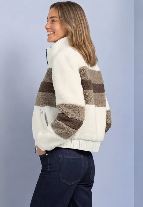 Junge Frau in cremeweißer Teddy-Jacke mit braunen Streifen und Jeans lächelt seitlich vor hellem Hintergrund.