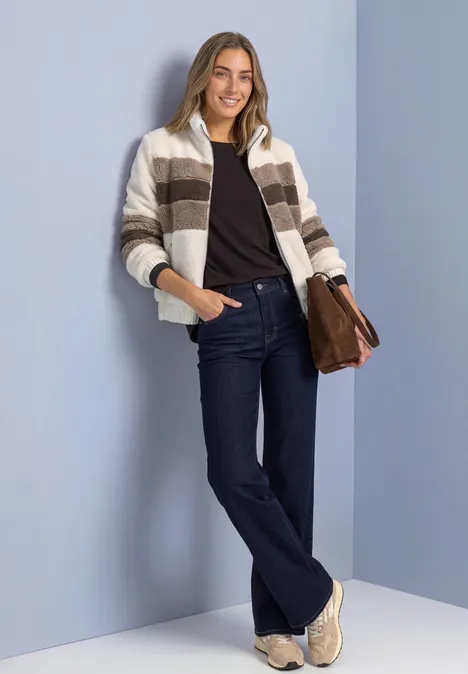 Frau mit gestreiftem Cardigan, dunkler Jeans, Sneakern und Tasche lehnt lächelnd vor lila Wand.