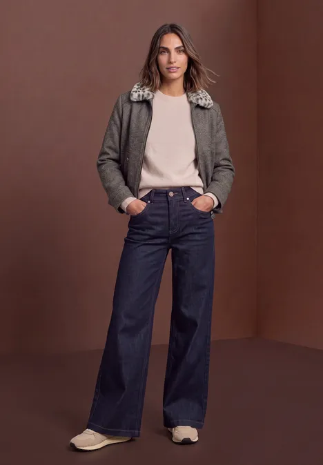 Frau in olivgrüner Jacke mit Pelzkragen, beigem Pullover, weiten Jeans und Sneakern vor braunem Hintergrund.