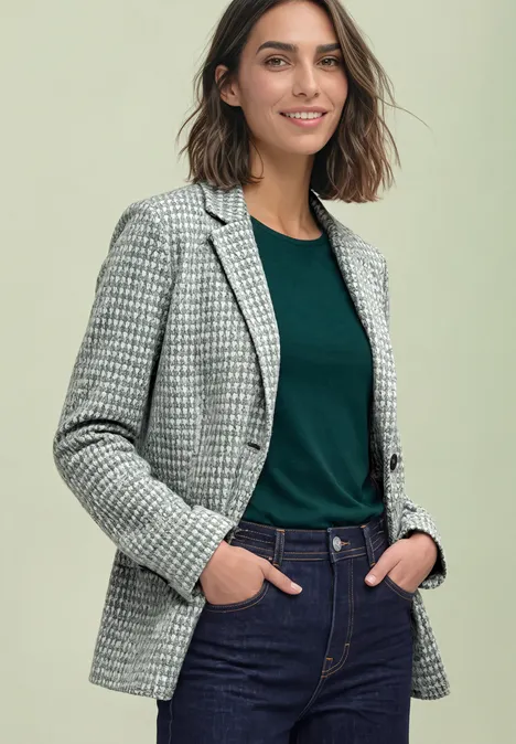 Frau trägt Hahnentritt-Blazer, grünes Shirt und Jeans, steht mit Händen in den Taschen und lächelt.