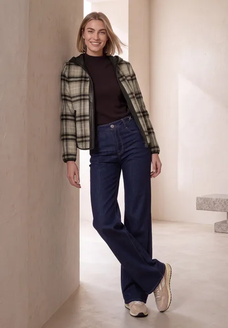 Modell in minimalistischer Umgebung lehnt lässig: karierte Jacke, schwarzer Pullover, weite blaue Jeans, weiße Sneaker.