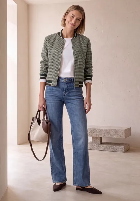 Blouson mit Zipper und Karomuster sanded amber Frau in grüner Bomberjacke, weißem T-Shirt, Jeans und Loafers trägt Tasche in minimalistischem Ambiente.