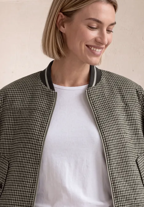 Blouson mit Zipper und Karomuster sanded amber Eine lächelnde Frau trägt eine karierte Bomberjacke und ein weißes T-Shirt vor schlichtem Hintergrund.