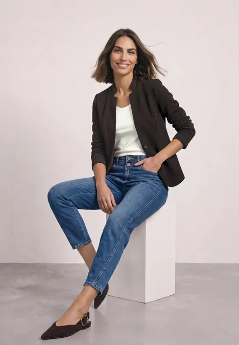 Eine Frau in Jeans, weißem Top und dunkelblauem Blazer sitzt lässig auf einem Würfel und lächelt.
