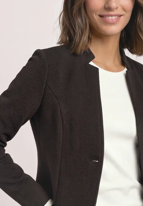Frau trägt dunkelbraunen Blazer über weißem Top, lächelt leicht.