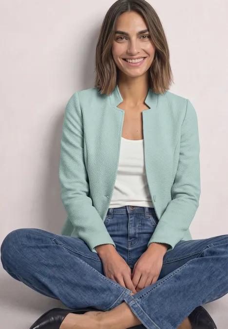 Eine lächelnde Frau sitzt im Schneidersitz und trägt ein hellgrünes Sakko, weißes Top und blaue Jeans.