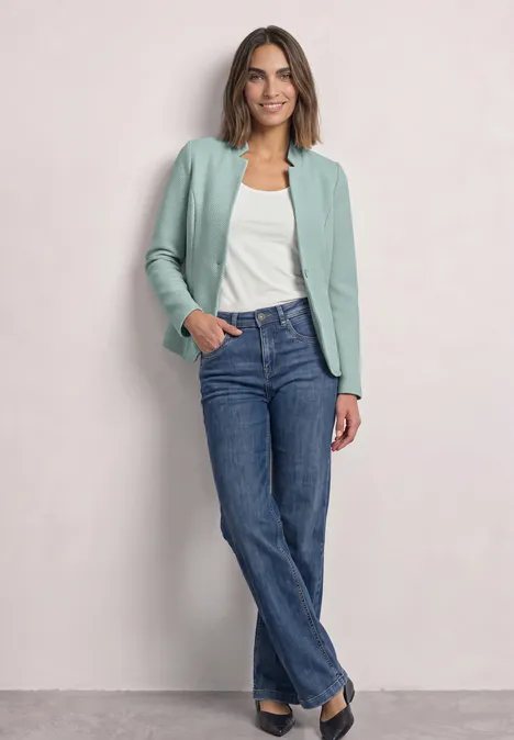 Frau trägt mintgrüne Jacke, weißes Top und blaue Jeans vor hellem Hintergrund.