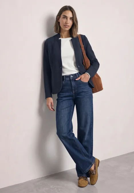 Vrouw in wit shirt, donkerblauwe blazer, rechte jeans, bruine mocassins en schoudertas tegen lichte achtergrond.